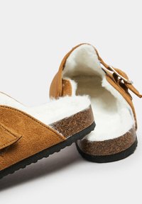 Zuecos de ante marrón con un forro blanco acolchado, plantilla de corcho y suela de goma negra. Presentan una hebilla lateral para ajustabilidad.