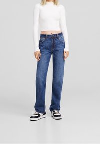 Blaue Jeans aus Denim mit lockerer Passform, fünf Taschen und kontrastierender Naht, kombiniert mit einem taillierten weißen Ripp-Crop-Top und schwarz-weißen Sneakers.