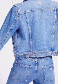 Light blue denim jacket with a cropped design, gold hardware accents, and logo patch at back. Paired with matching high-waisted jeans.