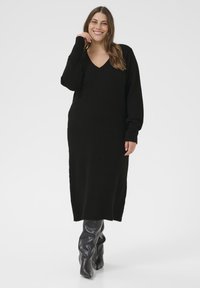 Schwarzes langärmliges V-Ausschnitt-Strickkleid aus strukturiertem, weichem Stoff. Das Modell trägt eng anliegende, knielange, glänzende schwarze Stiefel und steht vor einem weißen Hintergrund.