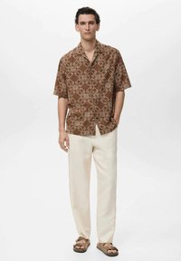 Braun kurzärmliges Hemd mit cremefarbenem abstraktem Muster, kombiniert mit cremefarbenen Hosen und braunen Sandalen. Einfacher, lässiger Schnitt und leichtes Material.