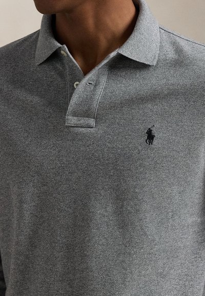 Muž nosi sivé polo tričko s dvoma gombíkmi a malým čiernym vyšívaným logom hráča pola na hrudi.