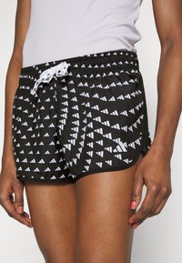 Shorts de sport noirs avec un motif triangulaire blanc et un cordon de serrage blanc portés par une personne à la peau foncée et au vernis corail.