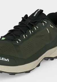 Σκούρο πράσινο και μαύρο παπούτσι για τρέξιμο σε μονοπάτια με μπεζ σόλα, μαύρα κορδόνια, υφασμάτινο επάνω μέρος και το όνομα της μάρκας τυπωμένο στο μπροστινό μέρος και στη γλώσσα.