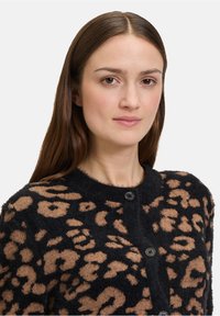 Hosszú barna hajú nő fekete kardigánt visel, amely bézs leopárdmintával díszített, közvetlenül a néző felé néz egy fehér háttér előtt.