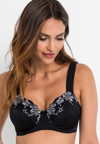 Reggiseno nero con motivi floreali ricamati e una parte superiore in pizzo trasparente, coppe modellate e spalline larghe per il supporto. Accento centrale a fiocco.