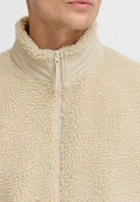 Beige fleece jacket with a high collar and zip closure. Features a soft, textured surface and a smooth collar section.