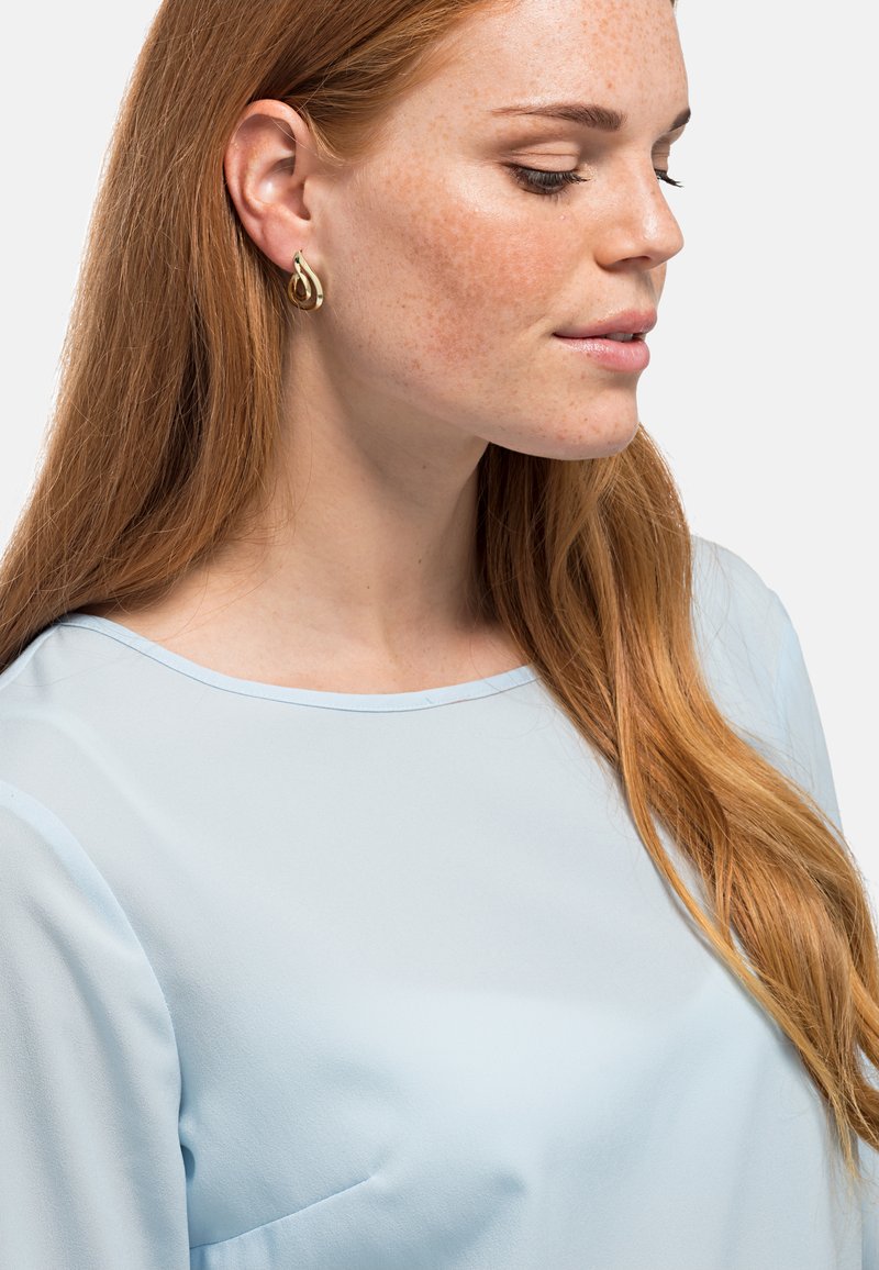 Gouden druppeloorbellen met zachte rondingen, gedragen met een lichtblauwe, vloeiende blouse met een ronde halslijn en zachte, doorschijnende mouwen.