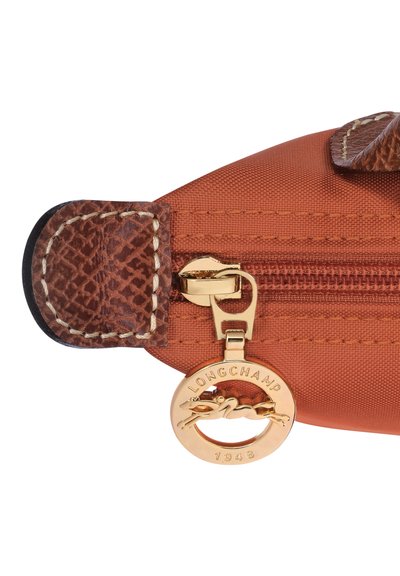 Orange textilebeutel mit braunem Lederakzent, goldfarbener Reißverschlussbeschläge und einem runden Logo-Anhänger mit der Inschrift "LONGCHAMP 1948."