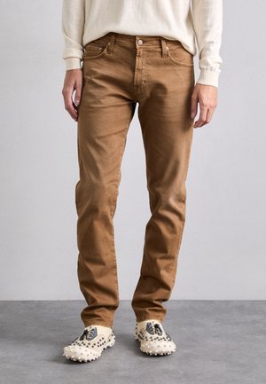 Jeans en coton marron avec une coupe ajustée et un design à cinq poches, dotés de quincaillerie métallique et d'un vieillissement minimal. Assortis avec des chaussures beige et grises.