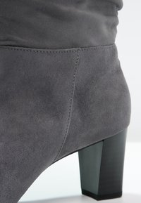 Gros plan sur une botte cheville en suède gris à talon bloc, avec coutures visibles et texture lisse sur fond neutre.