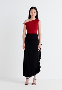 La modella sta in posa con sicurezza indossando un top rosso a spalla unica e una gonna nera con dettagli arricciati, su uno sfondo bianco semplice.
