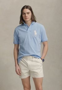 Light blue polo shirt with a two-button placket and embroidered logo on the left chest, paired with white shorts. Smooth cotton fabric.
