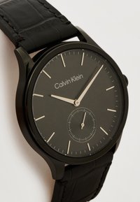 Μαύρο ρολόι χειρός Calvin Klein με μινιμαλιστικό καντράν, λεπτούς μπεζ δείκτες ώρας και δείκτες, ρυθμισμένο στις 9:05, με μικρό υποκαντράν δευτερολέπτων στις 6 η ώρα.