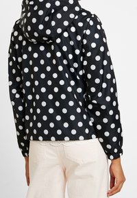 Černá bunda s bílými puntíky, vybavená kapucí, elastickými manžetami a zkráceným střihem. Látka vypadá hladká a lehká.