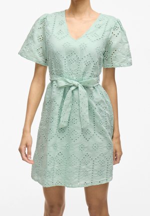 Femme portant une robe courte à manches courtes vert clair avec un motif en dentelle ajourée, un col en V et une ceinture nouée à la taille.