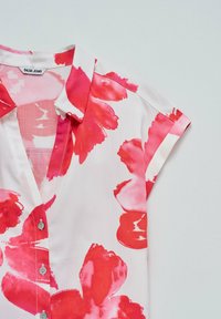 Blusa a maniche corte in tessuto leggero, con una base bianca e vivaci motivi floreali rosa, chiusura con bottoni e dettaglio del colletto.
