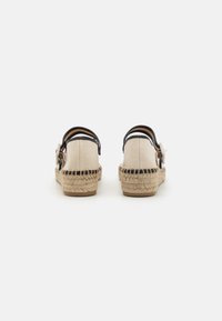 Beige espadrilleschoen met zwarte banden en een ronde knoopdetail. Jute-geweven zolen met textuurpatronen aan de zijkanten.