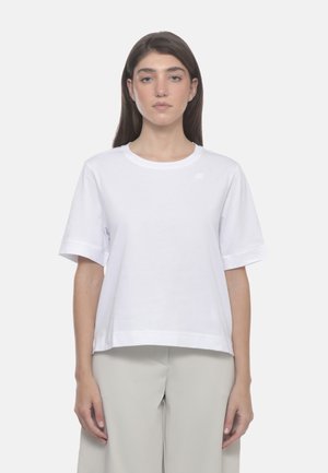 Donna con i capelli castani lunghi che indossa una maglietta bianca semplice a maniche corte e pantaloni beige chiaro, in piedi davanti a uno sfondo bianco.