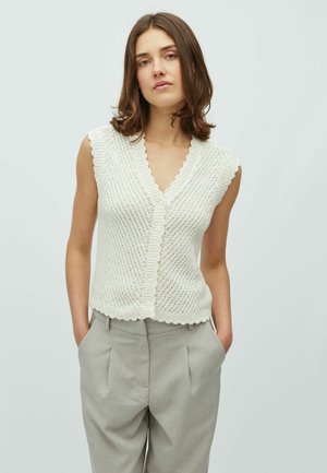 Bez rękawów, kremowy top dzianinowy z dekoltem w serek, ozdobiony falowanym brzegiem i zapięciem na guziki. W połączeniu z jasnoszarymi spodniami z wysokim stanem.