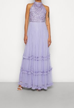 Femme portant une robe lavande sans manches à sequins avec un col halter et des volants superposés, associée à des sandales blanches à talons, debout sur un fond uni.