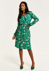 Green floral wrap dress with long sleeves and a tied waist, featuring various colorful flowers against a solid background. Stiletto heels.