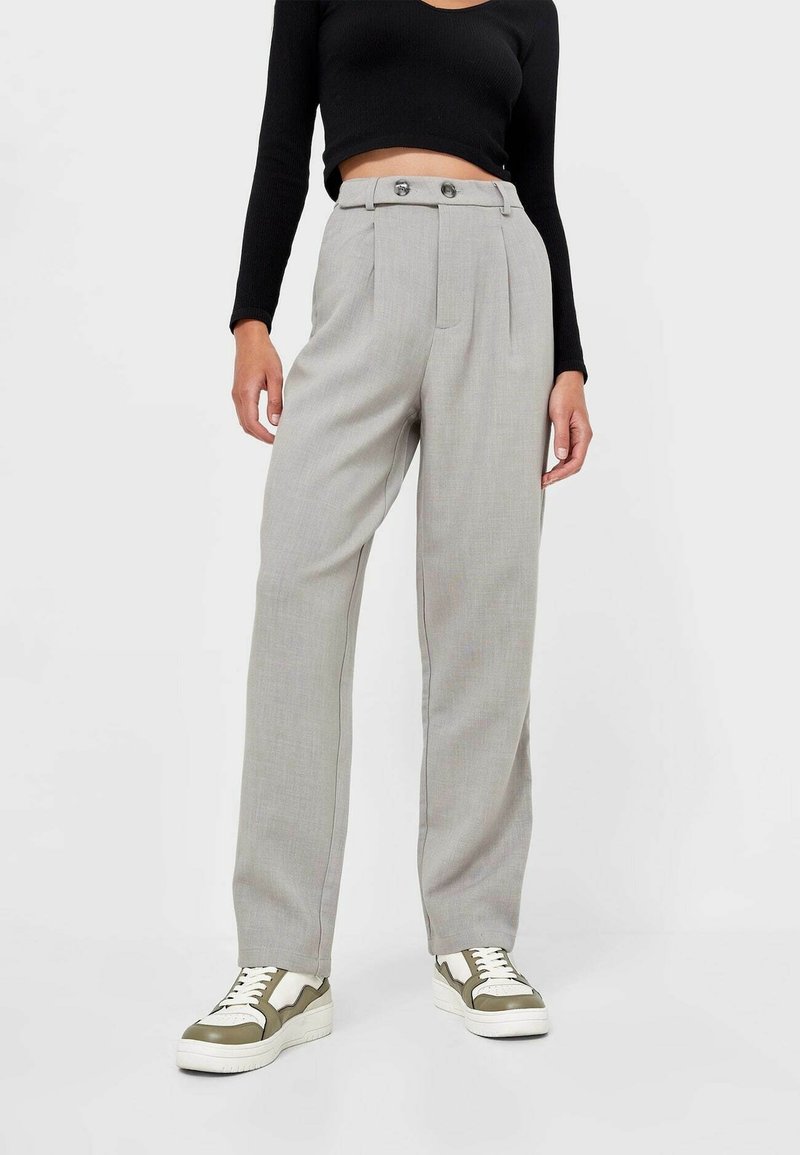 Pantalons ajustés gris clair avec deux boutons, taille haute et poches latérales, associés à des baskets vert olive et blanches.