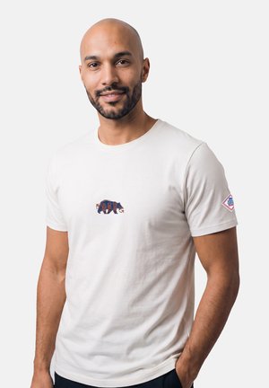 Entspannter Mann mit Bart, trägt ein weißes T-Shirt mit kleinem Bärendesign auf der Brust und einem Aufnäher am Ärmel, Hand in der Tasche, heller Hintergrund.