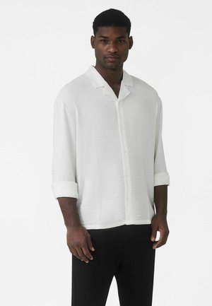 Cămașă albă cu nasturi, țesătură texturată și mâneci scurte suflecate, asortată cu pantaloni negri. Design simplu al gulerului, croială lejeră.