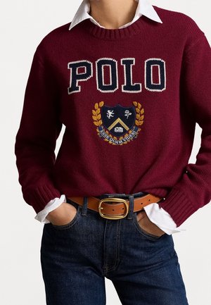 Personne portant un pull grenat avec "POLO" et un emblème de blason, une chemise blanche à col en dessous, une ceinture marron avec une boucle dorée et un jean bleu.
