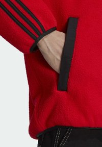 Chaqueta de forro polar roja con un acabado texturizado, acentos negros, tres franjas negras en las mangas y bolsillos laterales con bordes en contraste.