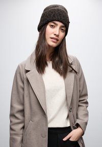 Gorro de punto marrón con un patrón texturizado, abrigo beige con solapas con muesca y suéter crema usado sobre pantalones oscuros.