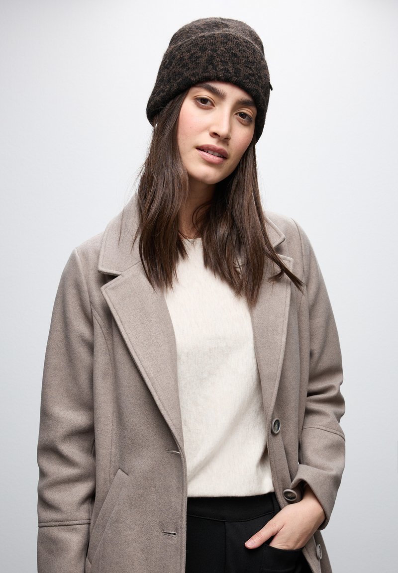 Gorro de punto marrón con un patrón texturizado, abrigo beige con solapas con muesca y suéter crema usado sobre pantalones oscuros.