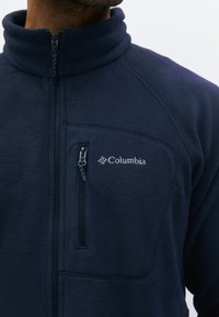 Muž nosící námořnicky modrou fleecovou bundu Columbia s kapsou na hrudi na zip a vysokým límcem.