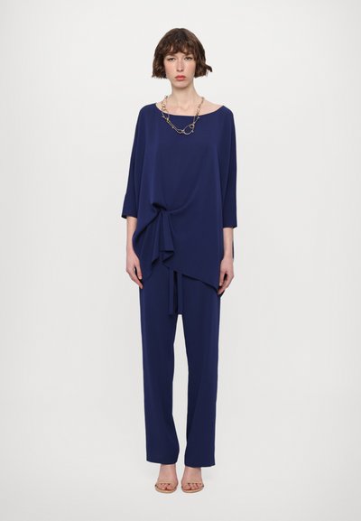 Navy blue loose-fitting top with a wide neckline and draped detail, paired with matching trousers. Features a chain necklace.