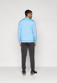 Jachetă sport de culoare albastru deschis, cu mâneci lungi și guler, purtată cu pantaloni gri închis și adidași negri cu accente verzi.