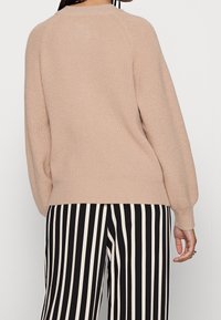 Light beige knitted sweater with ribbed texture, relaxed fit, and raglan sleeves. Worn with black and white striped pants.
