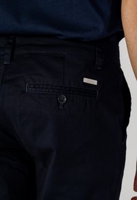 Pantaloni blu scuro realizzati in tessuto misto cotone. Presentano una tasca posteriore con bottone e una placca in metallo con logo. Texture liscia.