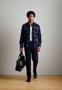 Chemise à carreaux bleu foncé sur un débardeur blanc, assortie à un jean sombre et des chaussures noires. Tient un sac en cuir noir.