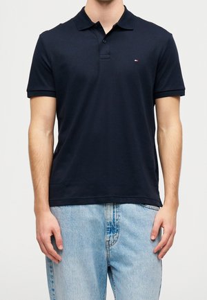 Mand iført en tætsiddende sort polo t-shirt med korte ærmer og lille logo på brystet, kombineret med lyseblå jeans mod en ensartet baggrund.