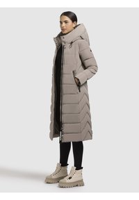 Cappotto lungo imbottito in beige chiaro con motivo a zigzag, caratterizzato da un alto colletto, due zippere laterali e dettagli hardware in contrasto.