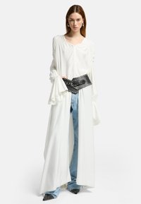 Weißes Langarmblusen-Shirt mit gerafften Schultern, kombiniert mit hellblauen weiten Jeans und einem schwarzen Nieten-Gürtel. Schwarze spitze Schuhe runden den Look ab.
