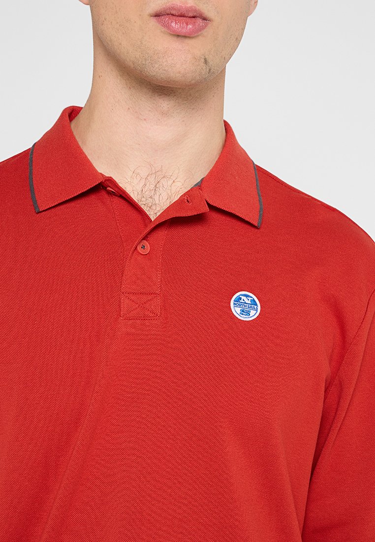 Hombre vistiendo una camiseta polo roja con un pequeño logo redondo de North Sails en el pecho, el cuello ligeramente abierto.
