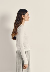 Žena s dugom smeđom kosom, odjevena u bijelu majicu s dugim rukavima i sive hlače, stoji u profilu na jednostavnoj bež pozadini.