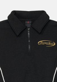 Pullover nero con colletto con zip, caratterizzato da un logo "Jordan" ricamato in oro e accenti laterali bianchi. Tessuto morbido con una consistenza liscia.