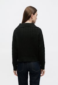 Černý krátký svetr s copánkovým vzorem, širokým límcem a texturovaným vzorem, vyrobený z měkkého pleteného materiálu. Nošený s tmavými džínami.