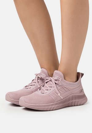 Par de zapatos deportivos de punto rosa con suelas estriadas, usados en pies desnudos sobre un fondo blanco.