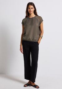 Top verde oliva con maniche corte e orlo arricciato, abbinato a pantaloni neri larghi. Il modello si trova su una superficie chiara, indossando sandali neri.