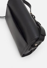 Černá kožená kabelka s cylindrickým tvarem, s hladkou strukturou, nastavitelním popruhem a stříbrnými kovovými detaily.