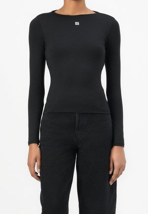 Kobieta ubrana w dopasowany czarny longsleeve z logo "HUGO" na piersi i czarne, wysokie spodnie dżinsowe na białym tle.